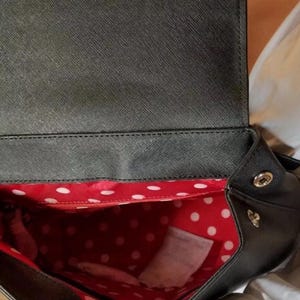 May include: Black backpack with a flap closure. The interior is red with white polka dots. The bag has a small gold-colored emblem on the side.