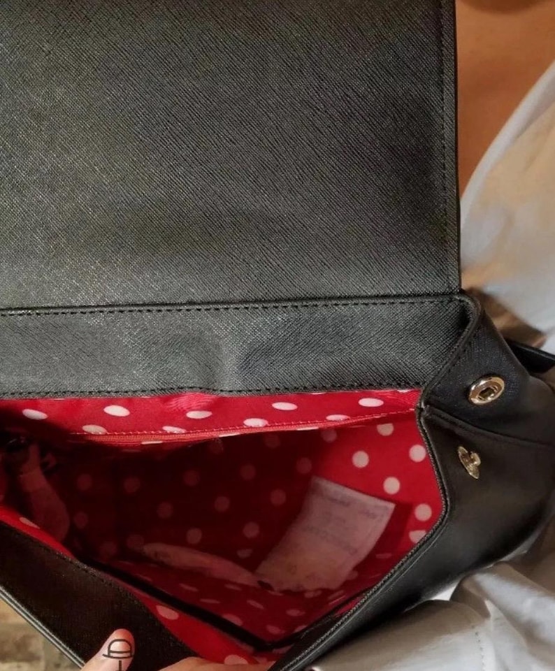May include: Black backpack with a flap closure. The interior is red with white polka dots. The bag has a small gold-colored emblem on the side.