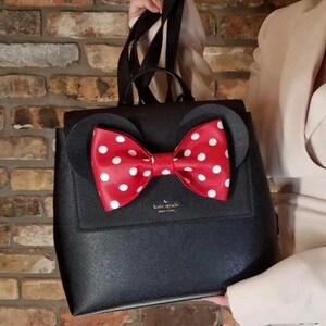 May include: Black backpack with a red bow featuring white polka dots. The backpack has two black circular ears and a flap closure. The brand name "kate spade" is visible on the front.