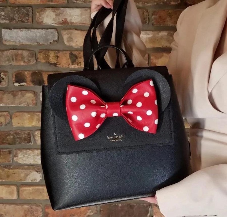 May include: Black backpack with a red bow featuring white polka dots. The backpack has two black circular ears and a flap closure. The brand name "kate spade" is visible on the front.