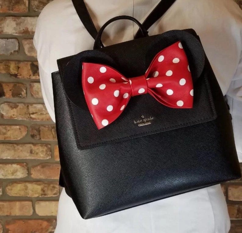 May include: Black backpack with a red bow featuring white polka dots. The bow is centered on the flap of the backpack. The backpack has a top handle and adjustable straps. The brand name "kate spade" is visible on the front.