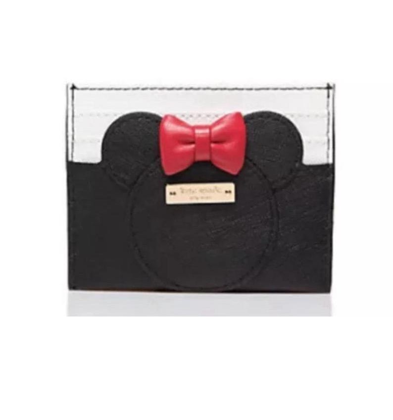 May include: Black and white cardholder with a Minnie Mouse design. The cardholder features a black face with two rounded ears and a red bow. A small gold rectangular tag is centered below the bow.