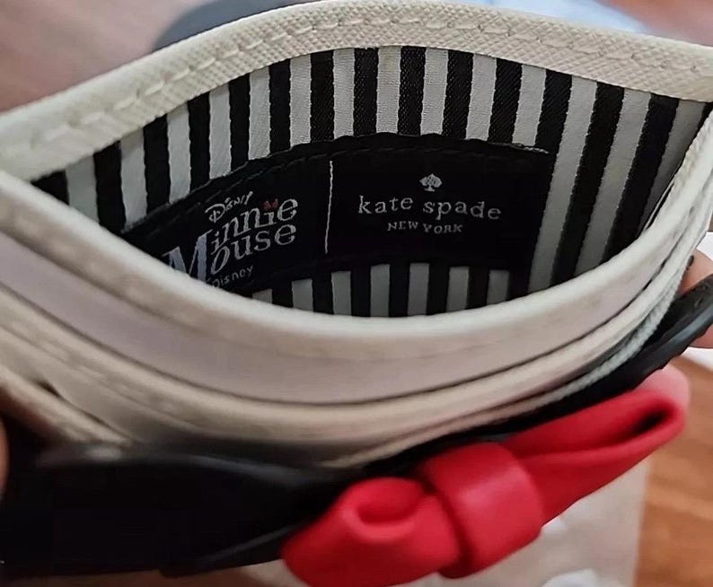 May include: A white and black cardholder with a red bow. The interior features black and white stripes and two labels: "Minnie Mouse Disney" and "kate spade NEW YORK".
