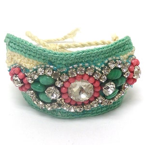 May include: A handcrafted bracelet featuring a turquoise crochet base with cream-colored accents. The bracelet is adorned with pink beads, green stones, and sparkling rhinestones. The bracelet is secured with a cream-colored string.