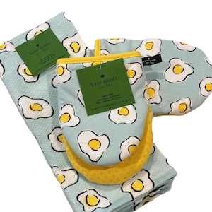 May include: A kitchen set featuring a light blue towel, oven mitt, and pot holder, all adorned with a fried egg pattern. The oven mitt and pot holder have yellow accents. The set includes tags from Kate Spade New York.