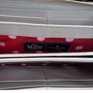 May include: A light gray wallet with a zipper closure. The interior features a red polka dot lining and a black label with "Disney Minnie Mouse" and "kate spade NEW YORK" branding.