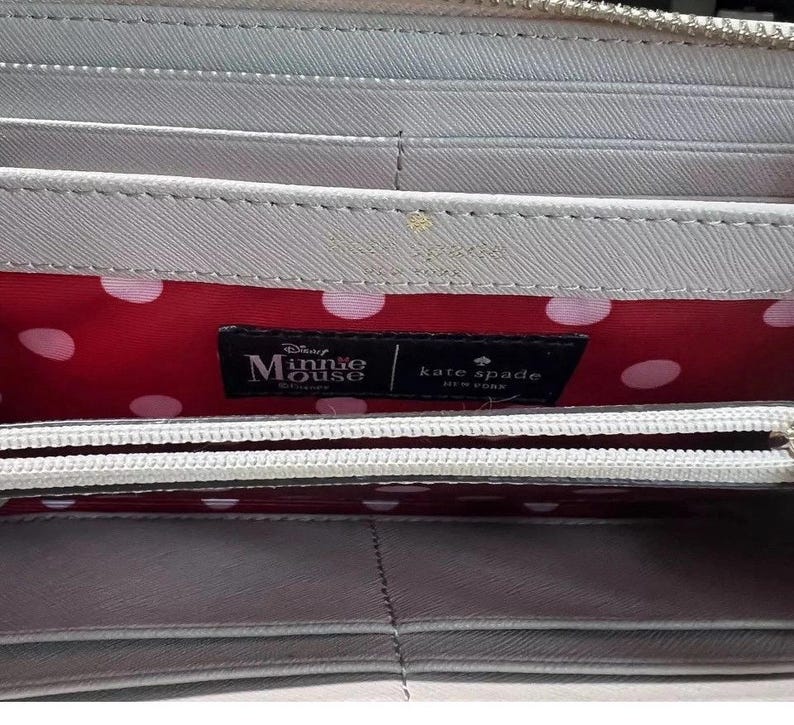 May include: A light gray wallet with a zipper closure. The interior features a red polka dot lining and a black label with "Disney Minnie Mouse" and "kate spade NEW YORK" branding.