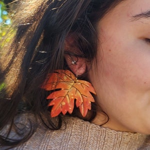 May include: Close-up of a person wearing a pair of autumn-themed leaf earrings. The earrings are shaped like maple leaves, featuring shades of orange, yellow, and red. The earrings are a fall fashion accessory.
