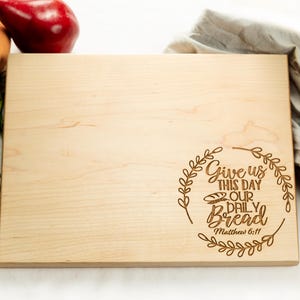 May include: A wooden cutting board with the text "Give us this day our daily bread Matthew 6:11" engraved in a circle with a wreath design.