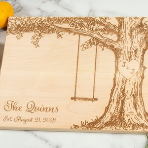 May include: A light-colored wooden cutting board with a tree and swing design. The text "The Quinns" and "Est. August 21, 2021" are engraved on the board. A heart with the initials "J + J" is also engraved.