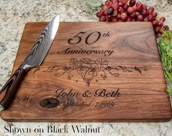 May include: A personalized wooden cutting board with the text "50th Anniversary John & Beth Established 1969" engraved on it. The board is shown on a black walnut surface.