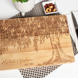 May include: Personalized wooden cutting board with a laser-engraved weeping willow tree design. The board features the text "Michael & Stephanie est. 2023" and the initials "M & S" carved into the tree trunk.