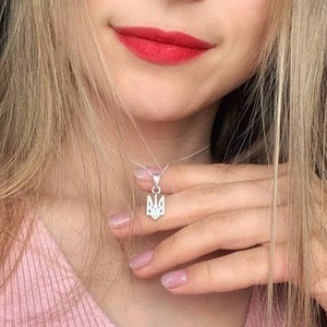 Puede incluir: Un collar con colgante de plata con el emblema ucraniano Tryzub. El colgante cuelga de una fina cadena. La persona lleva una camiseta rosa, tiene el pelo largo y rubio y lápiz labial rojo.