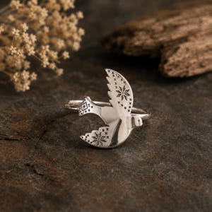 Op de afbeelding: Een zilveren ring met een vogel in vlucht als middelpunt. De vogel heeft gegraveerde ster- en kruispatronen op de vleugels en staart. Het metaal heeft een gepolijste afwerking en de ringband is dun en rond, ontworpen voor om de vinger.