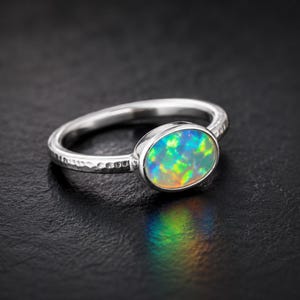 May include: A silver ring featuring a hammered band and an oval opal gemstone. The opal showcases a vibrant play of colour, including green, blue, and yellow hues. The ring is set against a dark background.