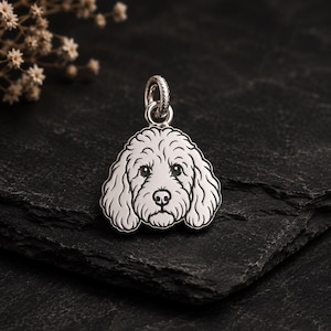 Puede incluir: Un colgante de metal plateado con forma de cabeza de perro de pelo rizado, que presenta detalles grabados de los ojos, la nariz y la textura del pelaje. El colgante incluye una pequeña anilla circular en la parte superior para una cadena.