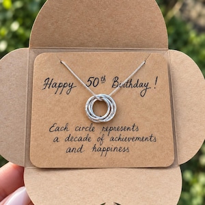 May include: A silver necklace featuring five interlocking rings suspended on a delicate chain, presented on a brown card with the text: Happy 50th Birthday! Each circle represents a decade of achievements and happiness. The jewelry is displayed on a card.