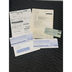 May include: A collection of various documents, including receipts, a memorandum with the Arista logo, and several forms. The documents are arranged on a black quilted surface. The papers are white with black text.