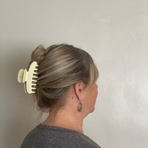Peut inclure: Une personne aux cheveux bruns portant une chemise grise et une grande pince &agrave; cheveux en plastique blanc cass&eacute;.