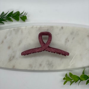 May include: A burgundy plastic hair claw clip with a textured finish. The clip is shaped like a heart and has a smooth, rounded edge.