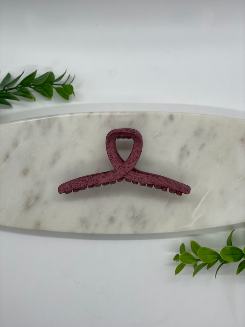 May include: A burgundy plastic hair claw clip with a textured finish. The clip is shaped like a heart and has a smooth, rounded edge.