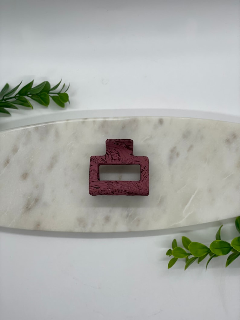 May include: A burgundy hair clip with a swirl pattern. The clip is rectangular with a rounded top and bottom.