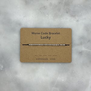 May include: A brown card with the text "Morse Code Bracelet Lucky" and "HANDMADE ITEM". A silver bracelet with small beads is displayed on the card.