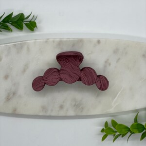May include: A burgundy plastic hair claw with a textured, swirl design. The claw is on a white marble surface.