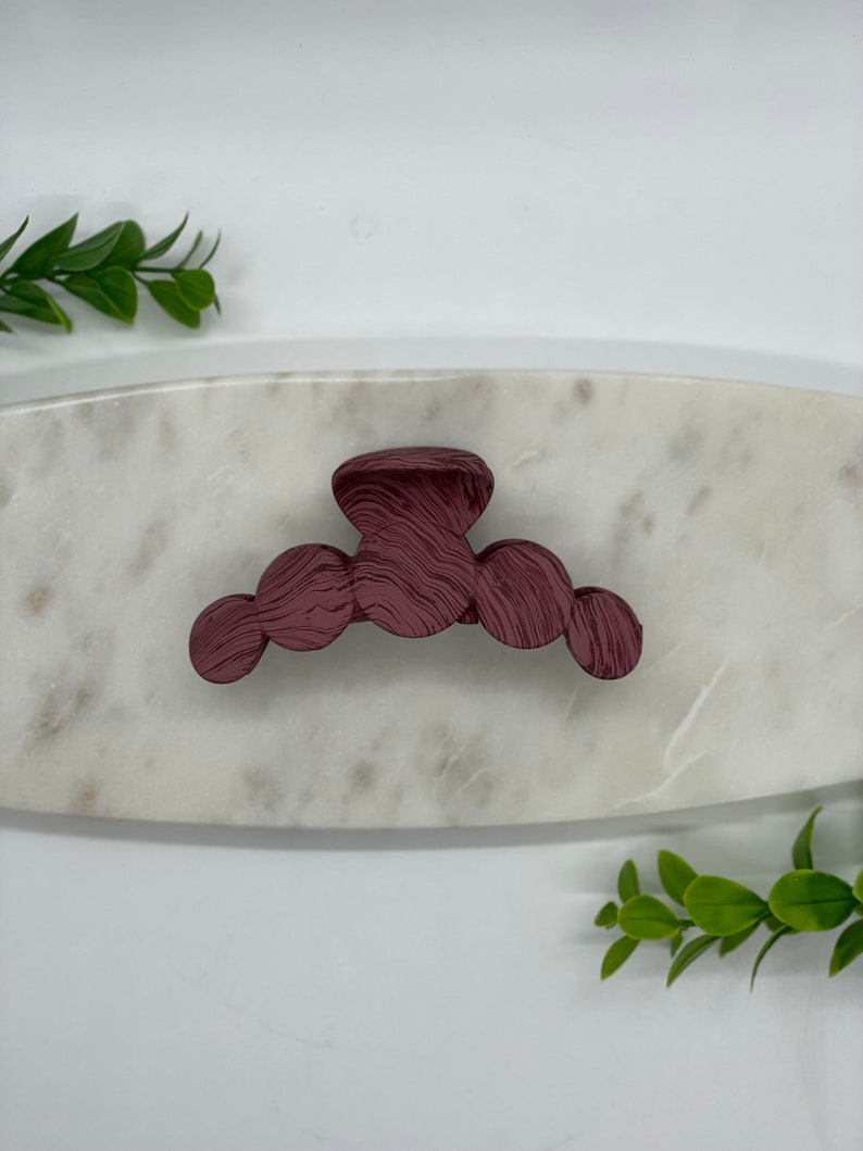 May include: A burgundy plastic hair claw with a textured, swirl design. The claw is on a white marble surface.