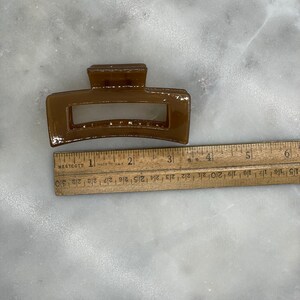 May include: A brown rectangular hair claw clip. The clip is approximately 3 inches long. The clip is made of a glossy material and has a rectangular opening in the center. A wooden ruler is next to the clip.
