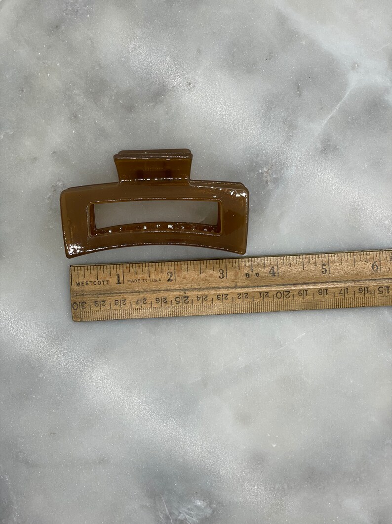 May include: A brown rectangular hair claw clip. The clip is approximately 3 inches long. The clip is made of a glossy material and has a rectangular opening in the center. A wooden ruler is next to the clip.
