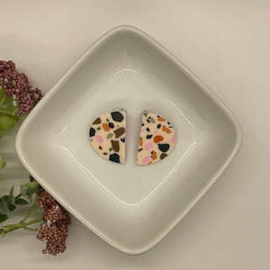 May include: A pair of pink and white half-circle earrings with a terrazzo pattern in black, brown, and orange.