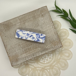 May include: A blue and white ceramic hair clip with a floral pattern. The clip is on a wooden surface with a white lace doily.