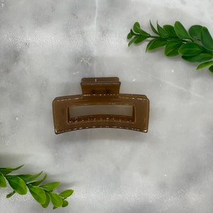 May include: A rectangular brown hair claw clip. The clip is made of a translucent material and has a simple, modern design. The clip is shown on a white and gray marble surface with green leaves.