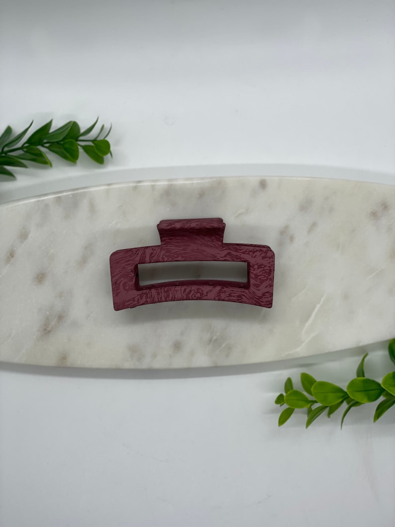 May include: A burgundy plastic hair claw clip. The clip is on a white marble surface.