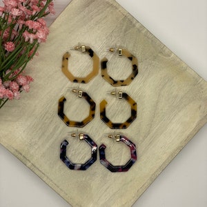 May include: Three pairs of geometric hoop earrings. The earrings are made of a tortoiseshell-like material with a gold-tone finish. The first pair is a light brown color, the second pair is a darker brown color, and the third pair is a blue and pink color.