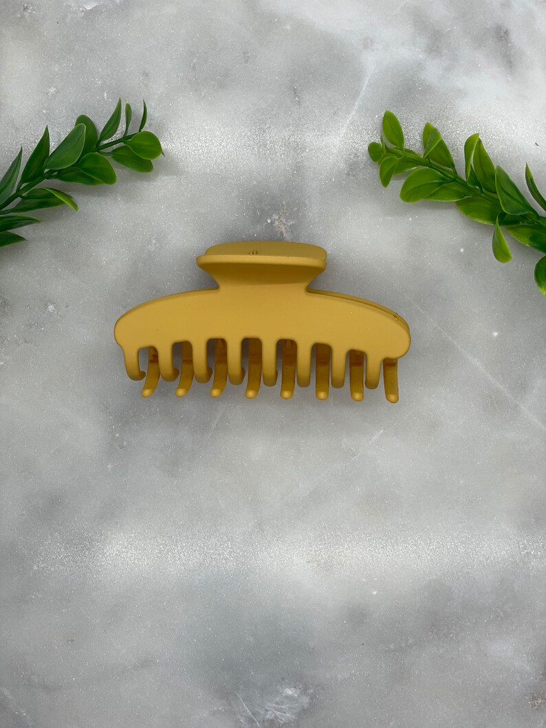 Peut inclure: Une pince &agrave; cheveux en plastique dor&eacute;e avec un dessus lisse et arrondi et plusieurs dents. La pince est sur une surface en marbre blanc.