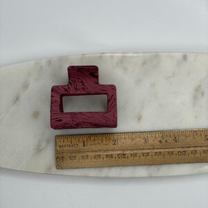 May include: A burgundy plastic hair clip with a marbled pattern. The clip is rectangular with a smaller rectangular opening in the center.