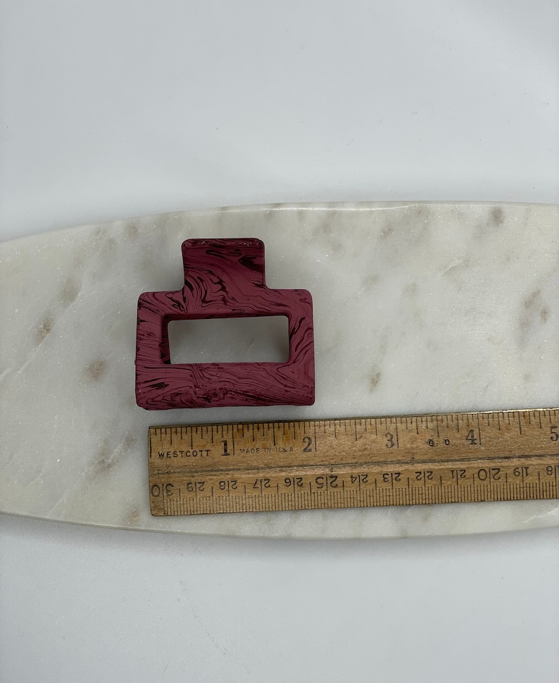 May include: A burgundy plastic hair clip with a marbled pattern. The clip is rectangular with a smaller rectangular opening in the center.