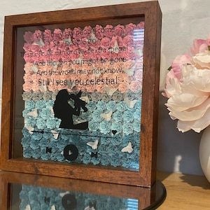 Puede incluir: Una caja de sombra de madera con un frente de vidrio lleno de flores de papel en tonos de rosa, azul y gris. Las flores están dispuestas en un patrón que se asemeja a un reproductor de música, con un botón de reproducción en el centro. El texto "Well, they say people come Say people go This particular diamond was extra special And though you might be gone And the world may not know Still, I see you celestial" está impreso en la parte superior de la caja.