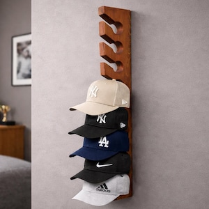 Peut inclure: Un porte-chapeaux en bois, fixé sur un mur gris, contenant cinq casquettes de baseball. Les casquettes sont de différentes couleurs, dont beige, noir et bleu marine, avec des logos de marques variés. Le support est de couleur marron foncé.