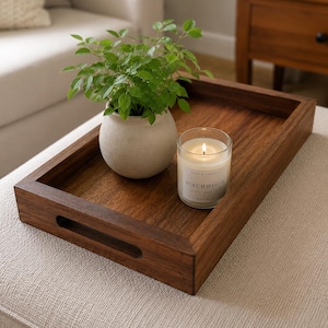 Peut inclure: Un plateau de service rectangulaire en bois brun foncé avec des poignées découpées. À l'intérieur du plateau se trouvent un petit pot en céramique blanc cassé contenant une plante verte et une bougie allumée dans un verre étiqueté Birchwood.