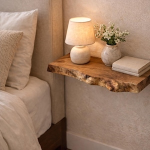 Peut inclure: Une étagère flottante en bois à bord brut fixée au mur. L'étagère supporte une petite lampe en céramique blanche avec un abat-jour en tissu, un vase en céramique blanche contenant des petites fleurs blanches et deux livres à couverture en tissu.