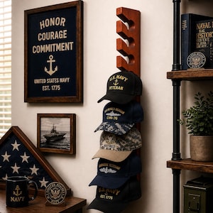Könnte beinhalten: Eine Präsentation mit einem hölzernen Hutständer mit sechs Baseballkappen im Marine-Stil. Ein gerahmtes Marine-Schild mit den Worten "Ehre, Mut, Engagement" und ein gerahmtes Foto eines Schiffes sind ebenfalls zu sehen. Eine Marine-Tasse und eine Holzflagge befinden sich auf einem Regal.