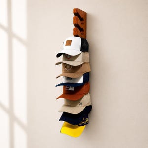 Op de afbeelding: Een houten hoedenrek met een verzameling baseballpetten. Het rek is aan een muur bevestigd en bevat zeven petten in verschillende kleuren, waaronder wit, beige, marineblauw en roest. De petten hebben verschillende logo's en ontwerpen.