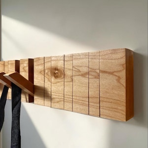Op de afbeelding: Houten wandkapstok met meerdere schuine haken. De kapstok is gemaakt van lichtgekleurd hout met zichtbare nerf en is aan een witte muur bevestigd. De haken houden zwarte items vast. Het ontwerp is minimalistisch en modern.