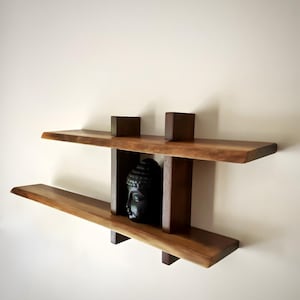 Puede incluir: Una estantería de madera con dos estantes y una escultura decorativa de la cabeza de Buda. Los estantes están hechos de madera natural con un acabado marrón oscuro. La unidad está montada en una pared blanca.