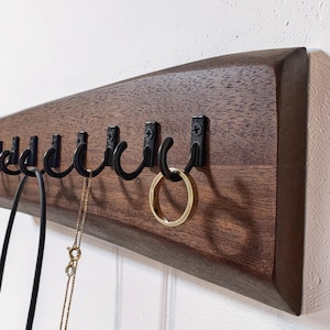 May include: A dark brown wooden wall-mounted hook rack with black metal hooks. A gold ring and a gold necklace hang from the hooks. The rack is mounted on a white wall.
