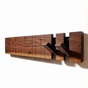 May include: A wooden wall-mounted coat rack with a dark brown finish. The rack features a series of vertical wooden slats and three projecting hooks. The hooks are attached to a black metal plate. The design is modern and minimalist.