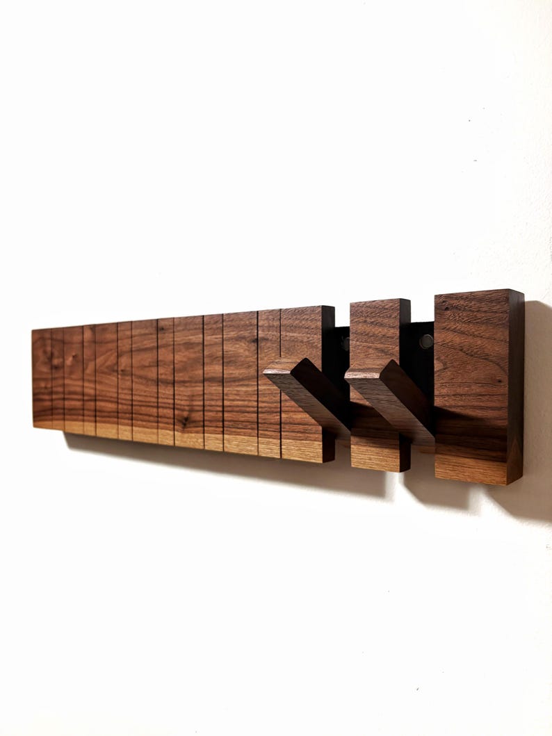 May include: A wooden wall-mounted coat rack with a dark brown finish. The rack features a series of vertical wooden slats and three projecting hooks. The hooks are attached to a black metal plate. The design is modern and minimalist.